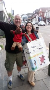 Man with Bearsac and Debra. Debra wearing home-made poster saying The bear is back in town. with her book cover and Bearsac underneath.