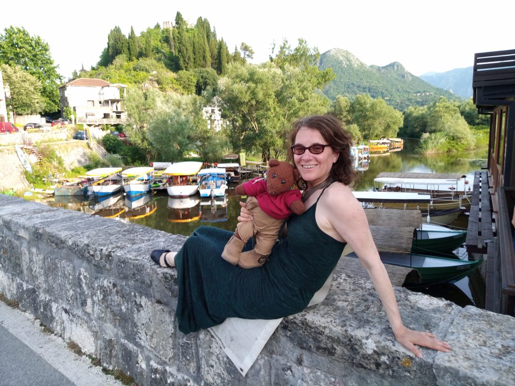 Debra sitting on bridge wall holding Bearsac. Lake and boat scenery.
