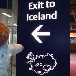 Bearsac the teddy bear in foreground of Exit to Iceland sign at te airport