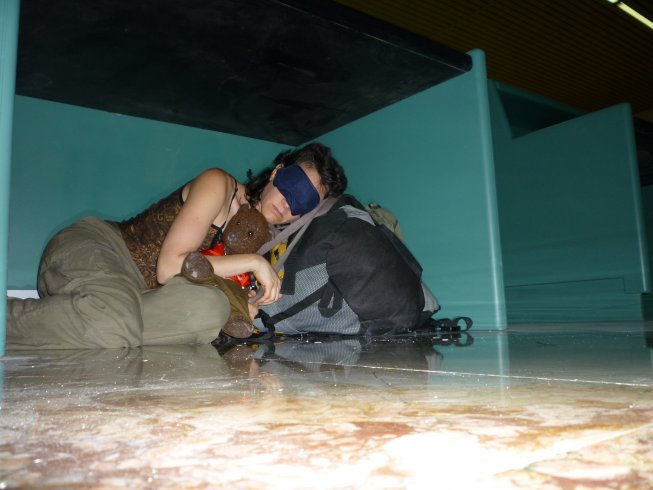Debra wearing an eyemask, cuddling Bearsac, whilst sleeping under an airport checkin desk
