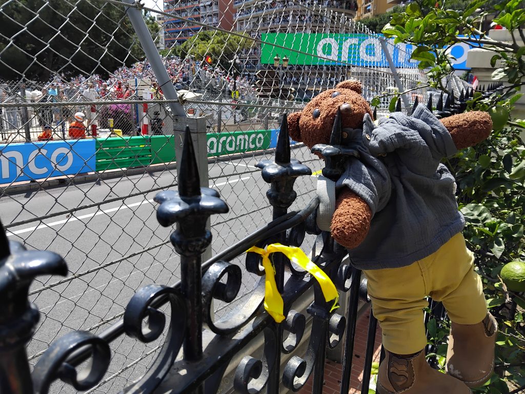 Bearsac the teddy bear in the foreground of the race-track with a yellow ribbon on the fence.