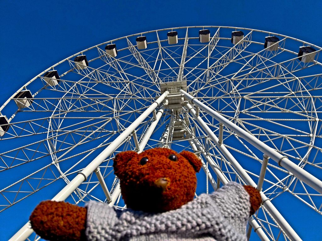 Bearsac the teddy bear in forground of a large ferris wheel.