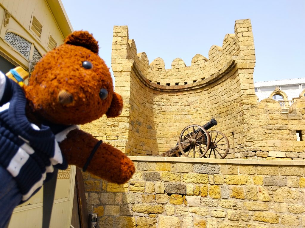 Bearsac the teddy bear in forground of a canon on a wall.