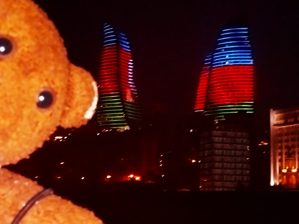 Bearsac the teddy bear in forground of the flame tower lit in blue red and green at night.