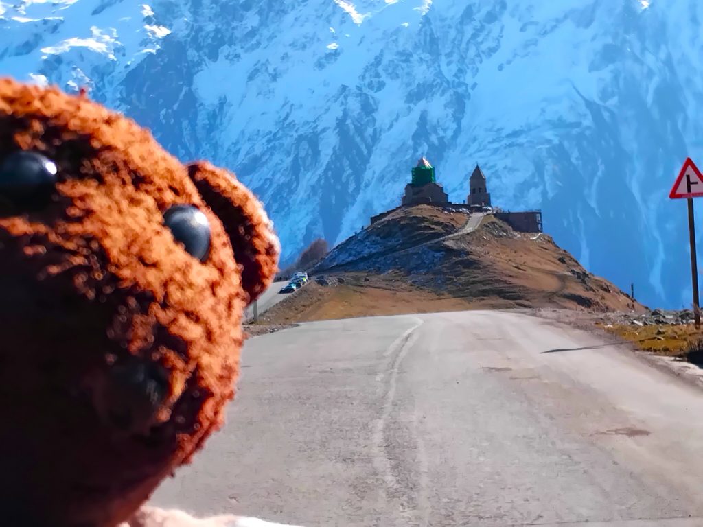 Bearsac the teddy bear in forground of Gergeti Trinity Church on the hill with snowy mountains behind.