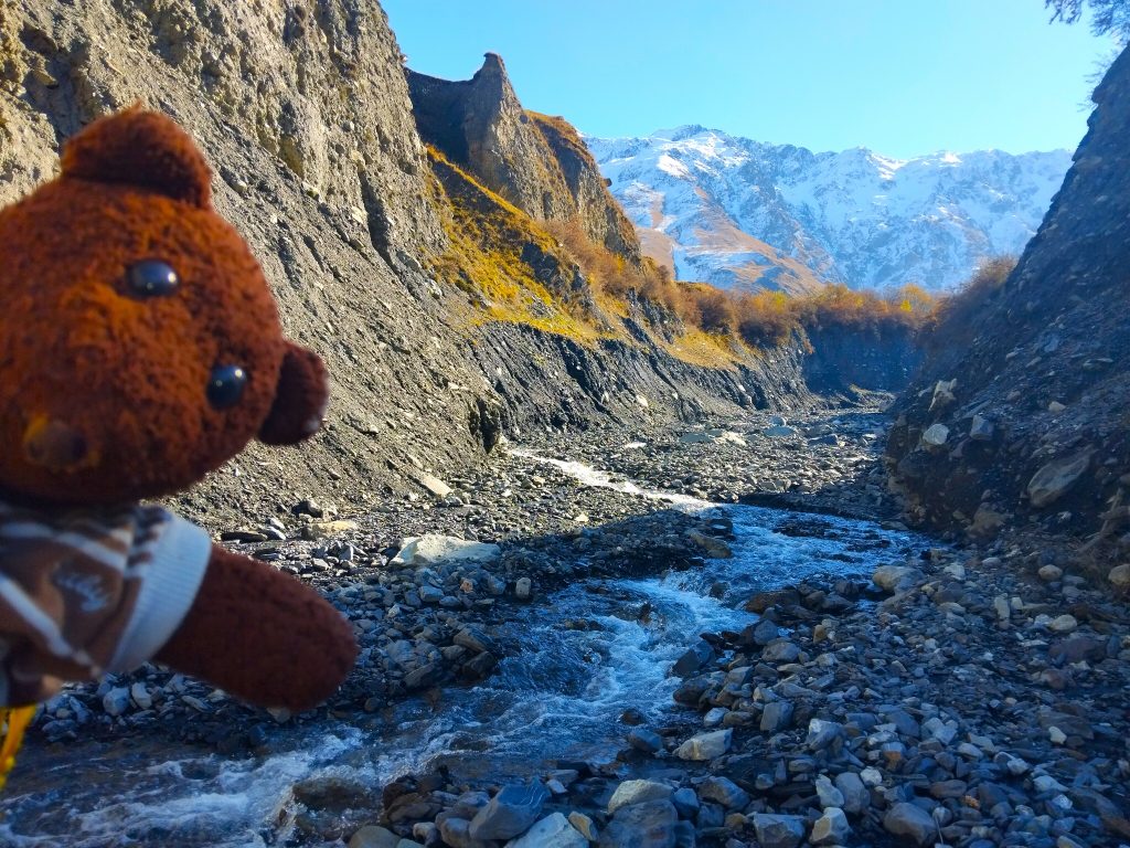 Bearsac the teddy bear in forground of a stony river and mountains view.
