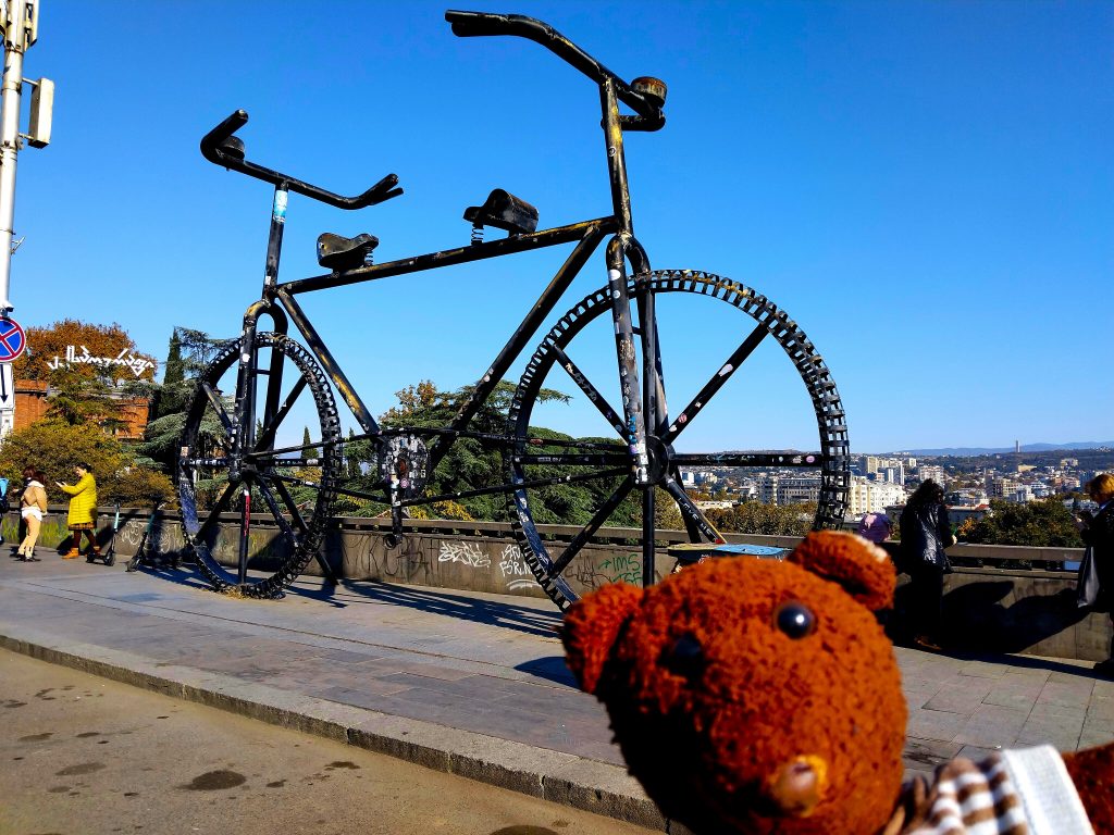 Bearsac the teddy bear in forground of a large bicycle monument and city view.