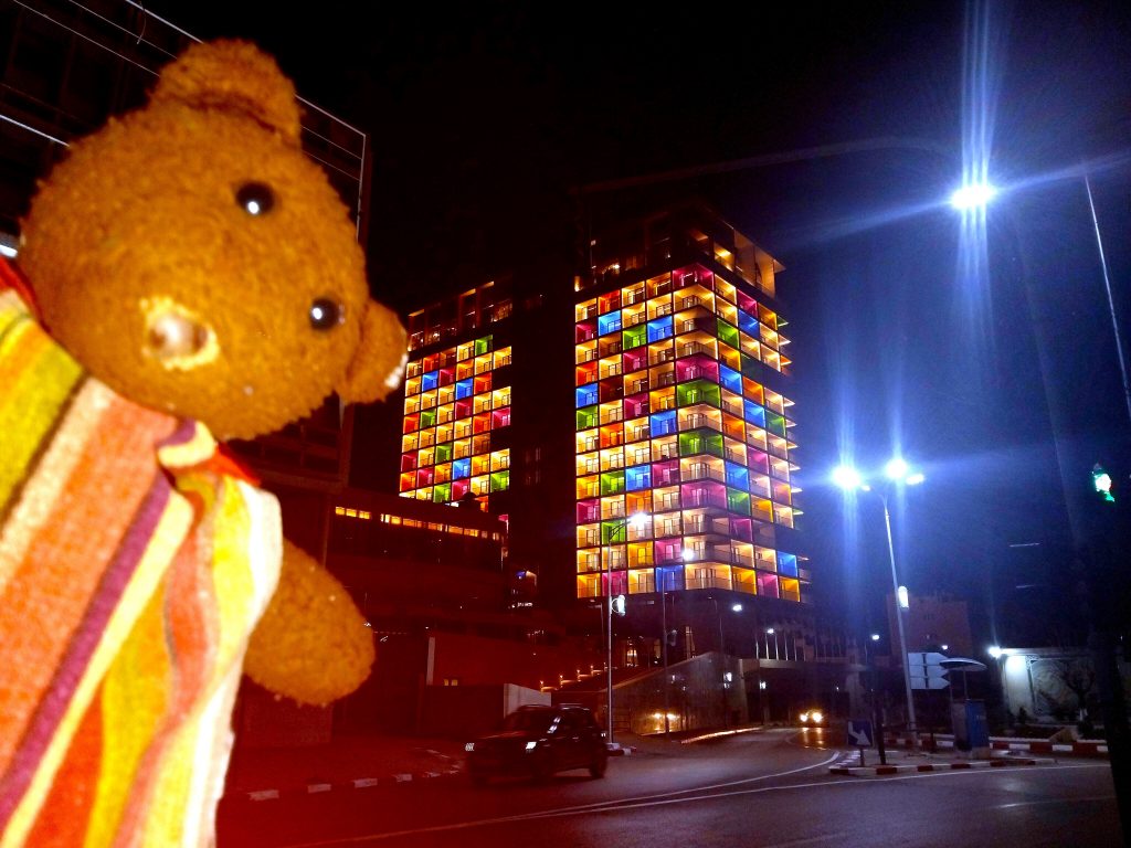Bearsac the teddy bear in forground of coloufukky lit building at night