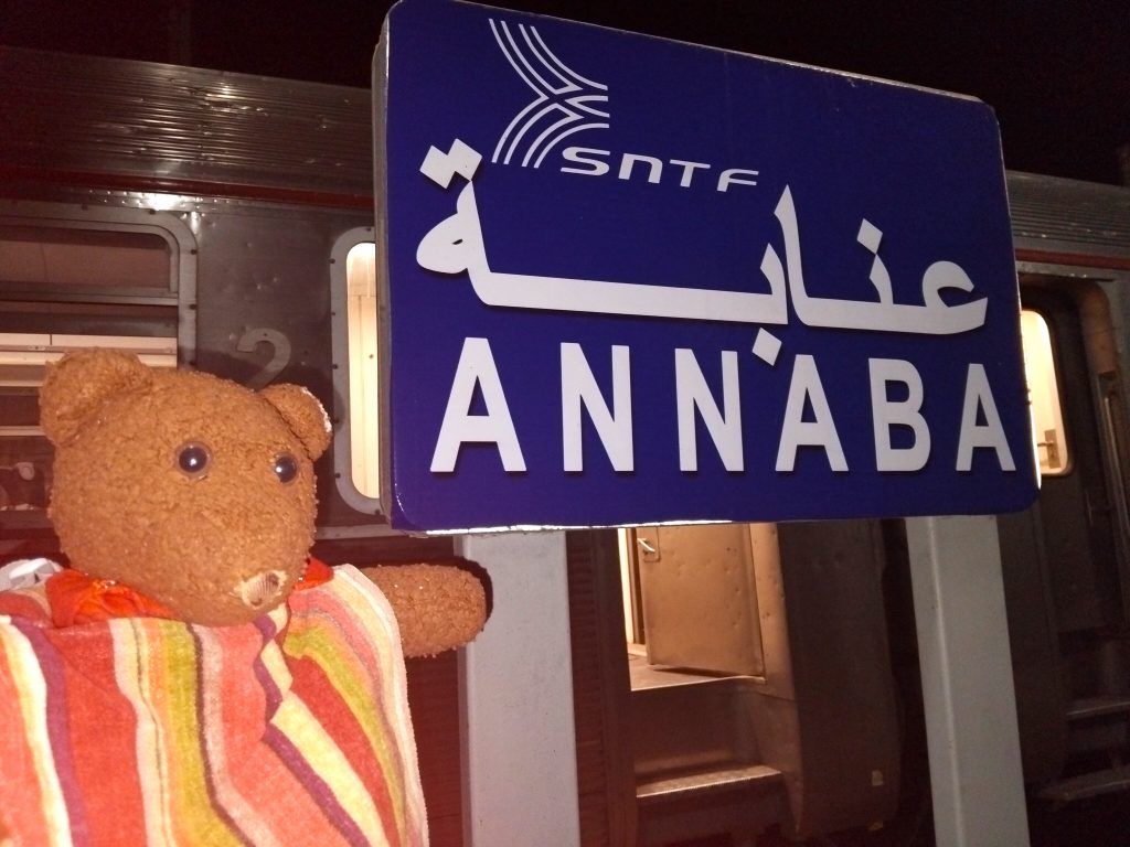 Bearsac the teddy bear in forground of a platform sign reading Annaba