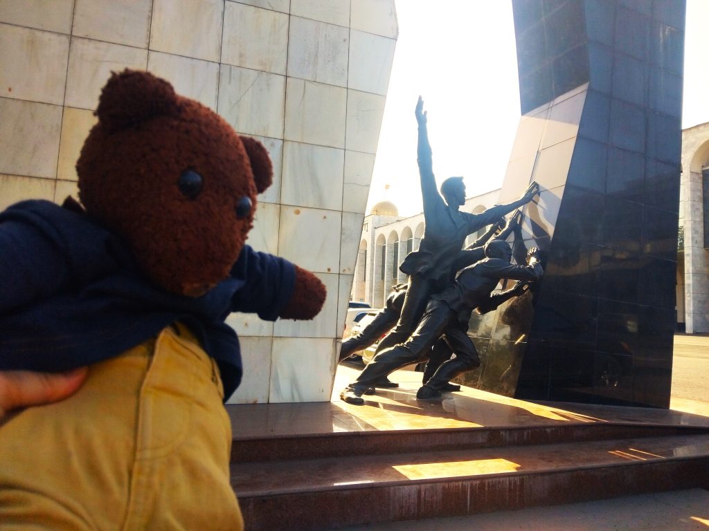 Bearsac the teddy bear in the forerground of a statue with men pushing a wall