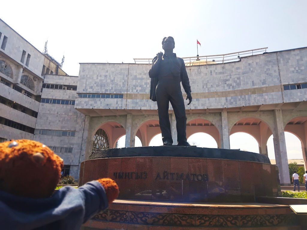 Bearsac the teddy bear in the forerground of a monument of Chingiz Aitmatov