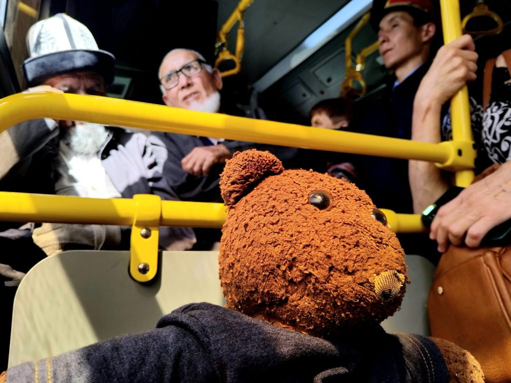 Bearsac the teddy bear in the forerground of huamns ona bus