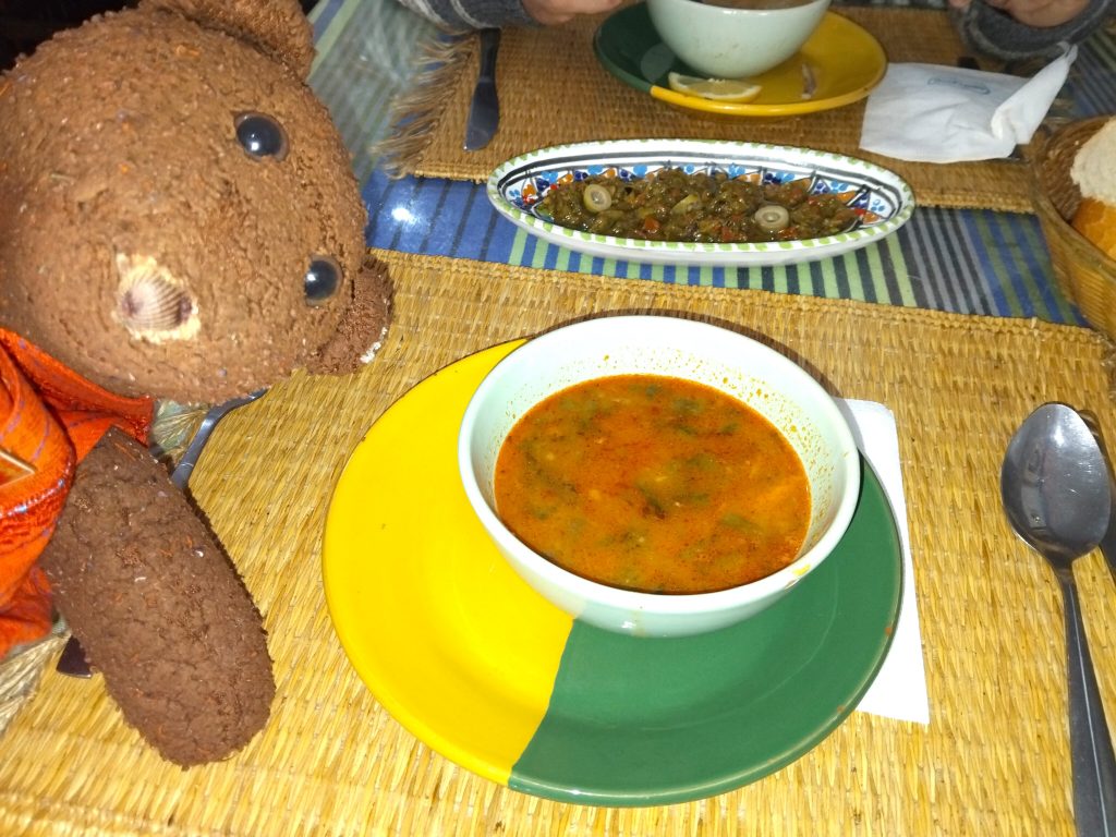 Bearsac the teddy bear peeping into the frame with a bowl of soup