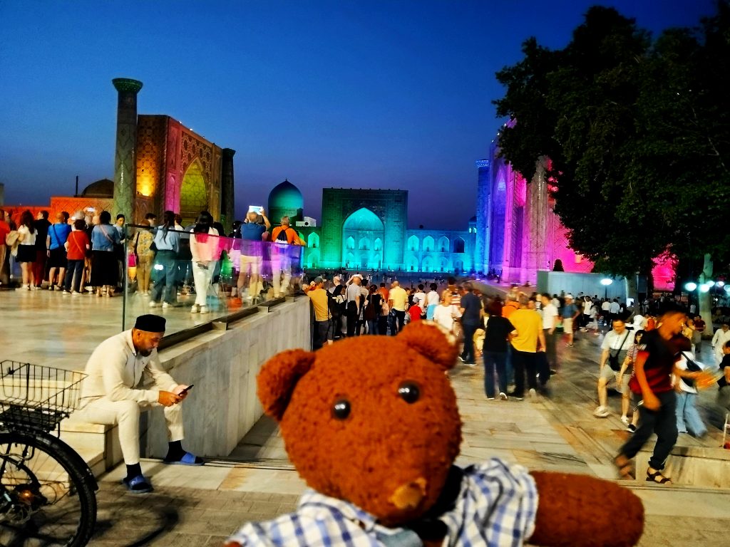 Bearsac the teddy bear in the foreground of amakand Registan Square with colorfully lit buildings