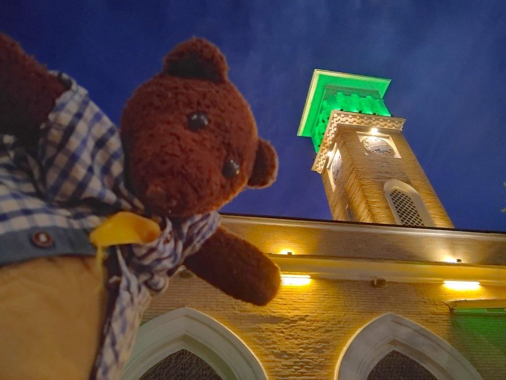 Bearsac the teddy bear in the foreground of a clocktower with green light.