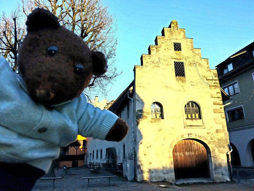 Bearsac the teddy bear in forground of a building with a stepped facet