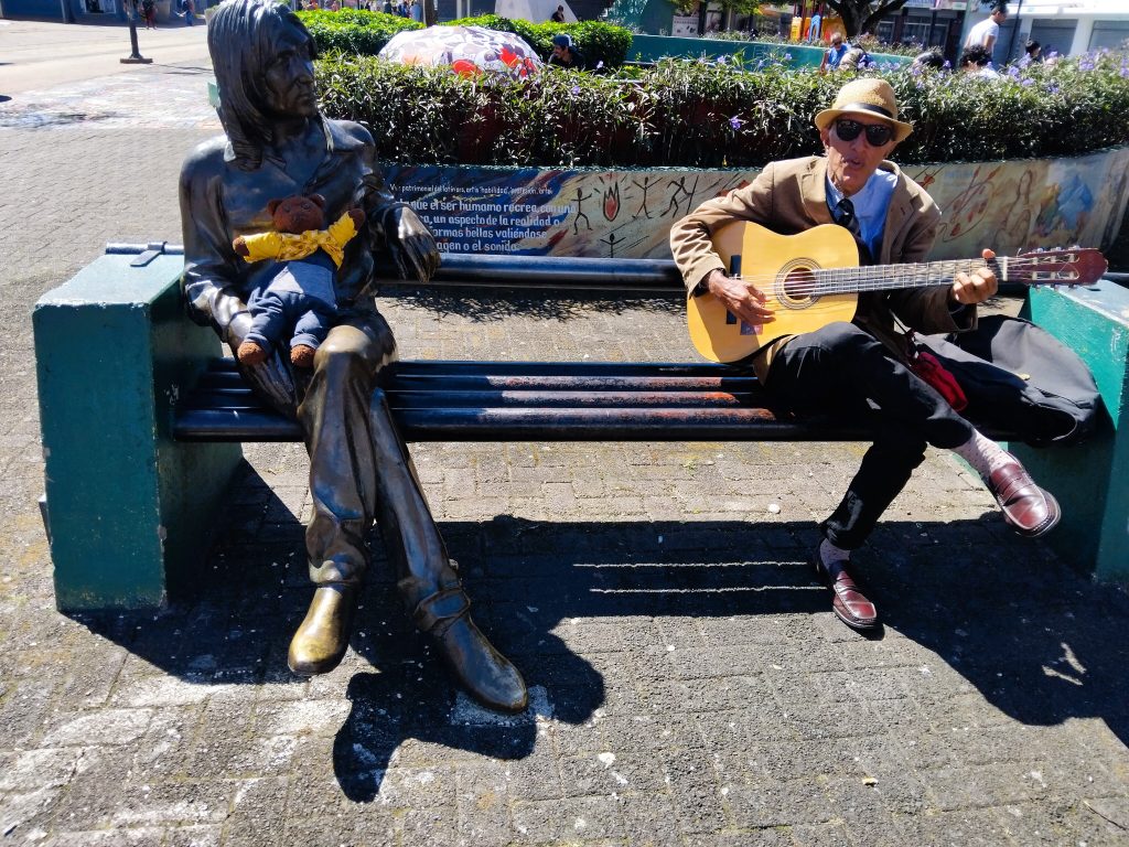Bearsac the teddy beaar sitting on a statue of Jhn Lennon next to a man playing a guitar.