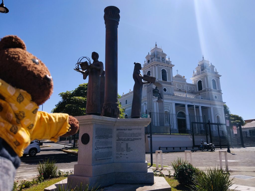Bearsac the teddy bear in foreground of a churh and monument.