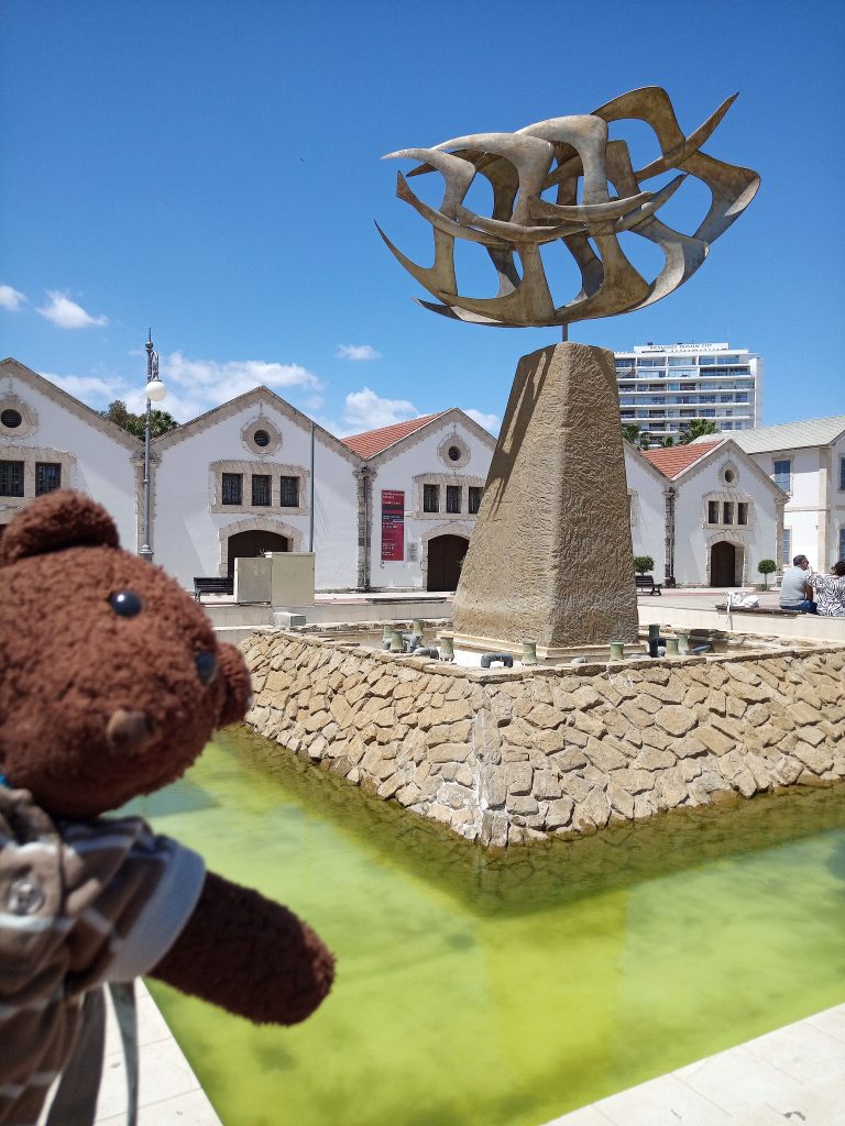 Bearsac the teddy bear in forground of the The Finikoudes Fountain, which is turned off.