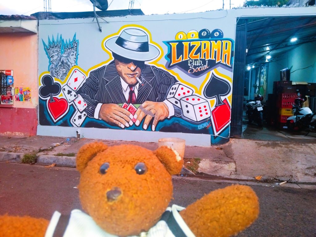 Bearsac the teddy bear in foreground Lizama Social Club murial of a suited man playing a card and dice game.