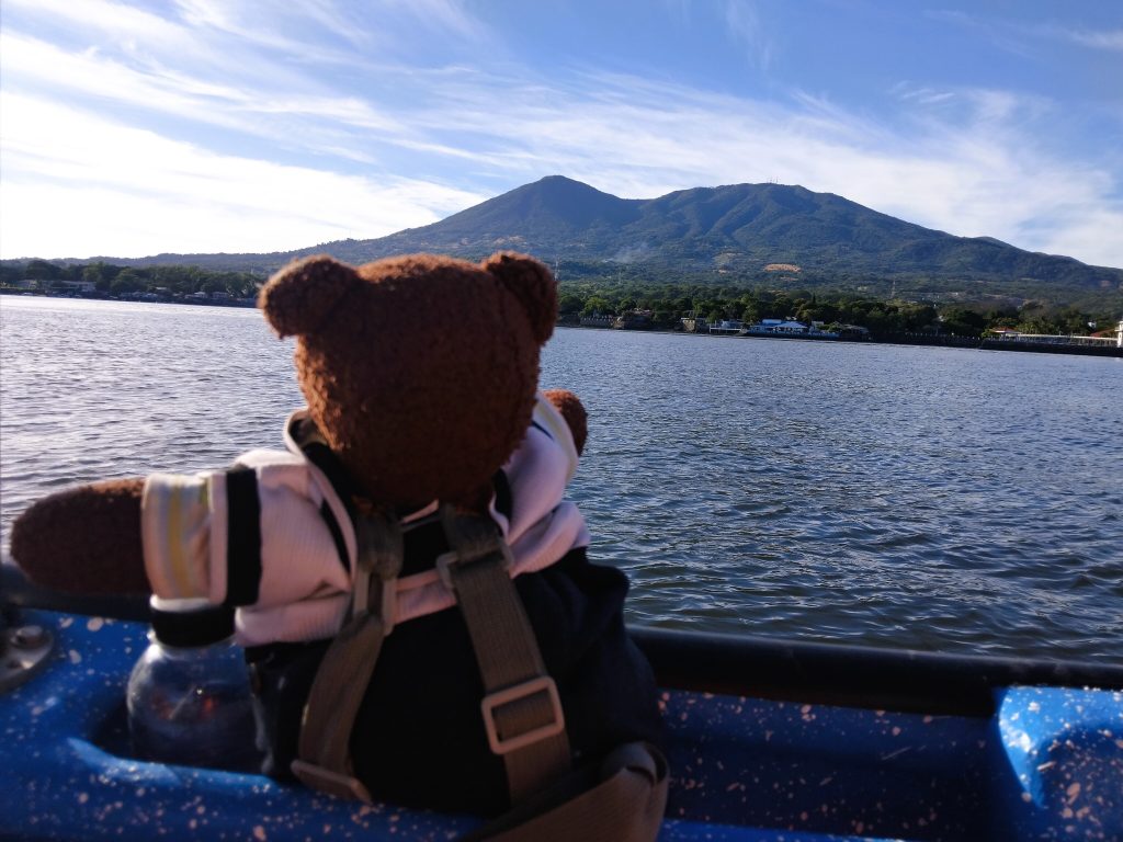 Bearsac the teddy bear on a small boat in foreground of the sea and mountains.