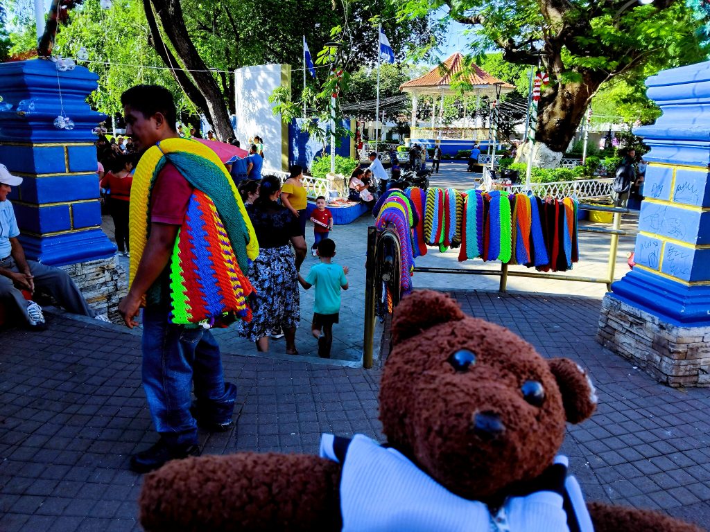 Bearsac the teddy bear in foreground of a man with brightly coloured hammocks slung over his shoulder and other slung over a railing.