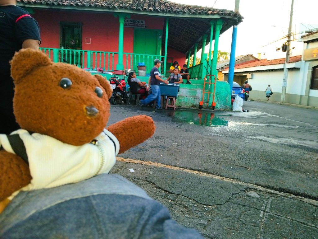 Bearsac the teddy bear in foreground of bright orange and green house with street hawkers.