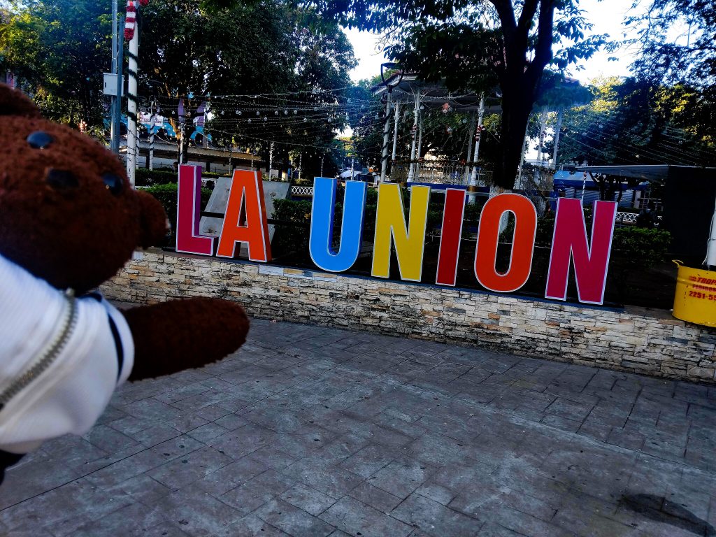 Bearsac the teddy bear in foreground of a text La Union monument.