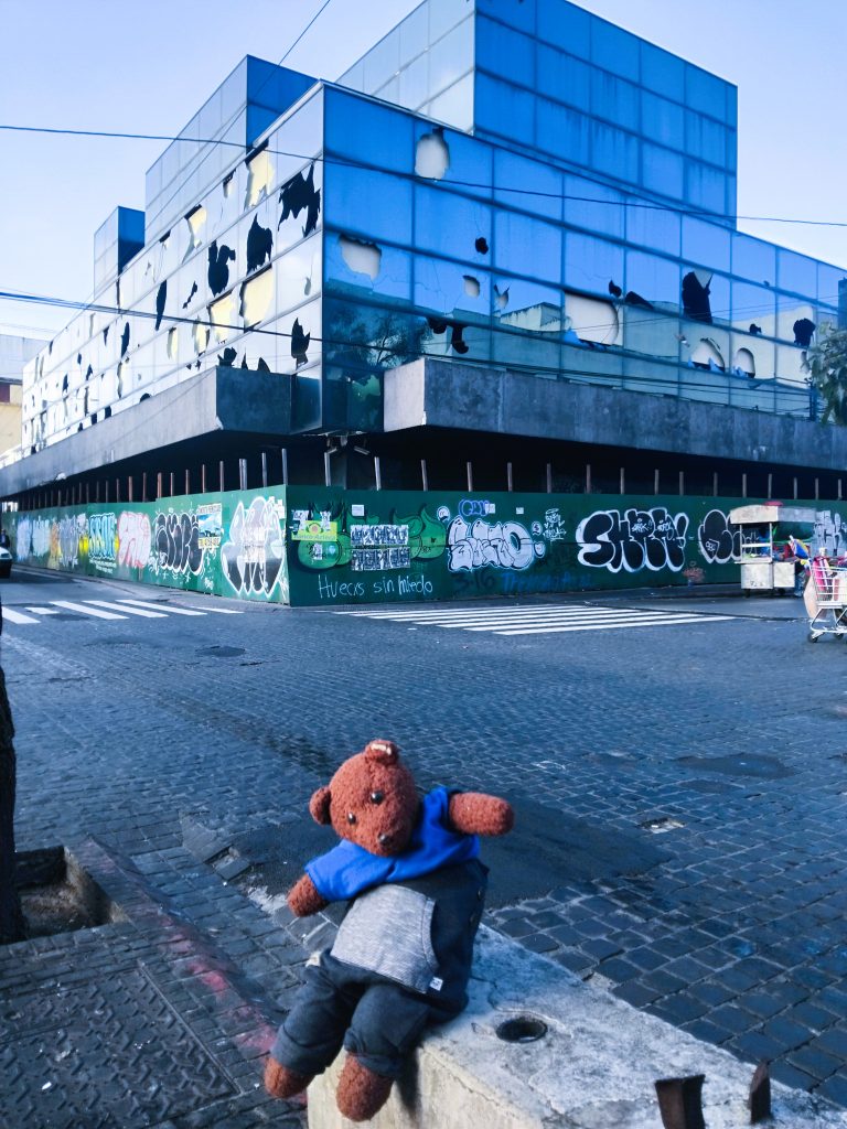 Bearsac the teddy bear in foreground of a glass building with many smashed windows.
