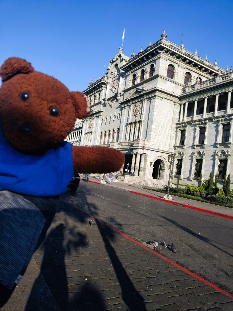 Bearsac the teddy bear in foreground of a grand building.