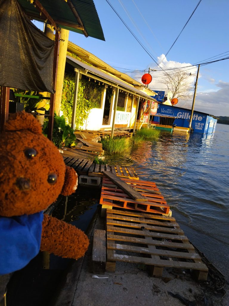 Bearsac the teddy bear in foreground of wodden shops with an overflown lake up to the entrances. and pallet boards as a walk-way.