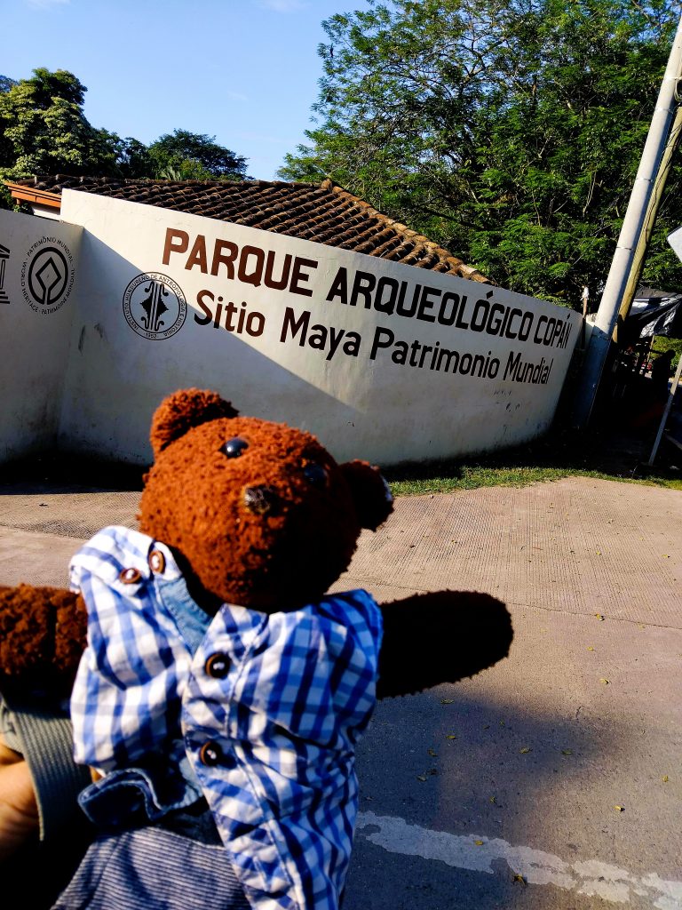 Bearsac the teddy bear in foreground of the entrance to Parque Arqueologico Copan.