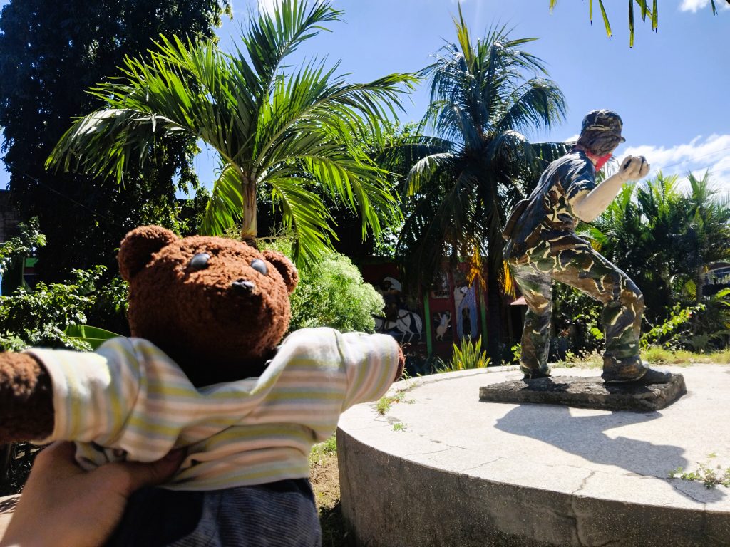 Bearsac the teddy bear in foreground of a museum piece of a solider throwing something.