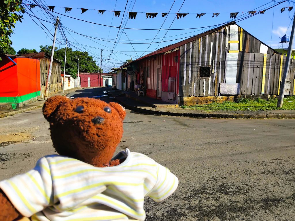 Bearsac the teddy bear in foreground of a weathered wooden house.