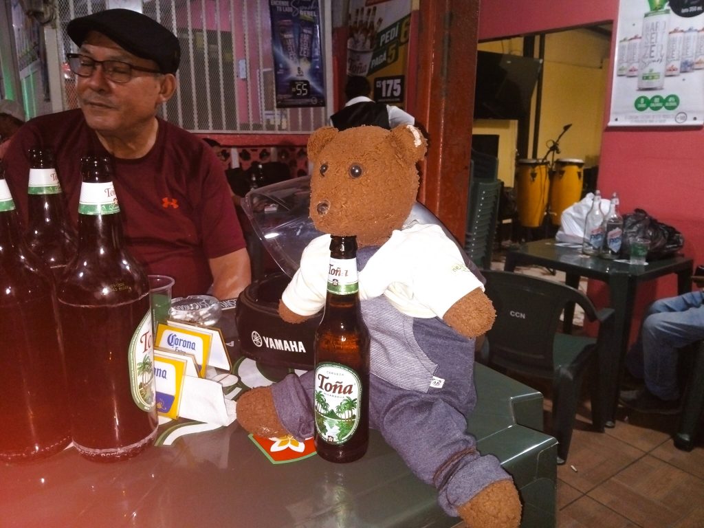 Bearsac the teddy bear with a bottle of beer under his chin. Sitting at a table witha man.