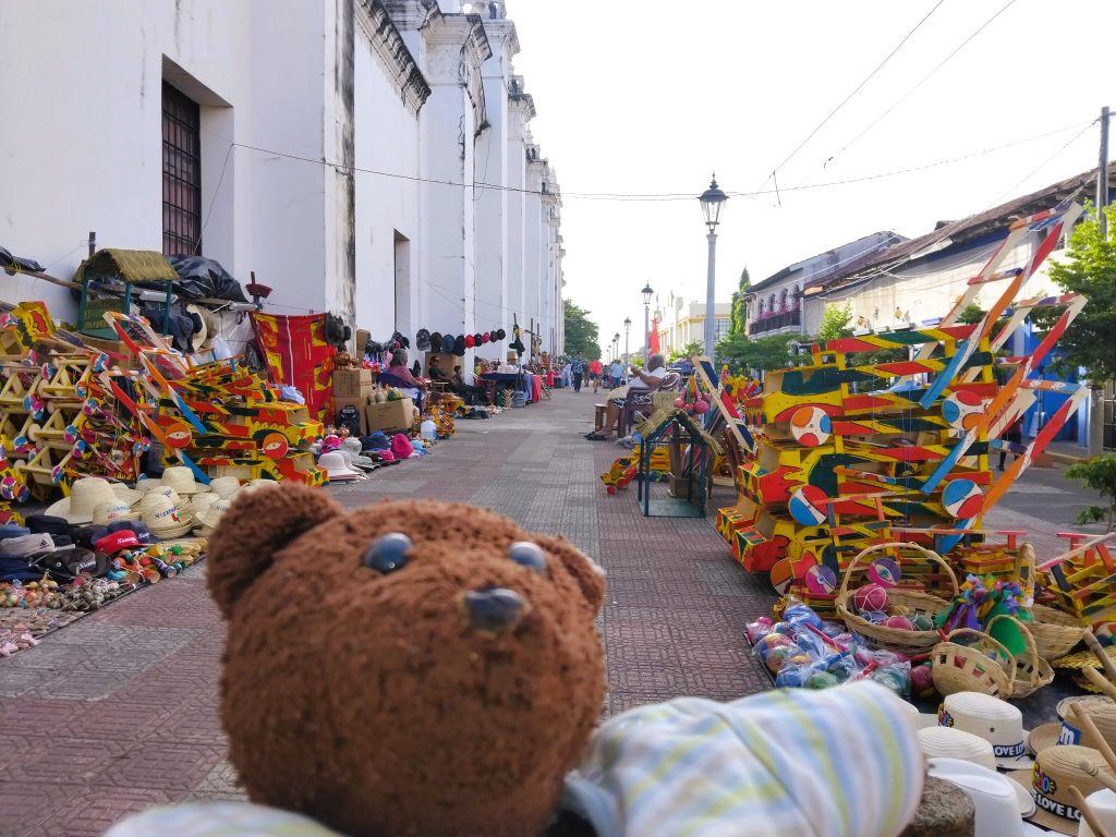 Bearsac the teddy bear in foreground of a market stall of colourfully paainted wooden items.