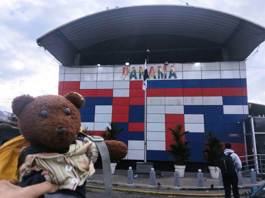 Bearsac the teddy bear in foreground of a Panana poassport control building, with the text. Panama.
