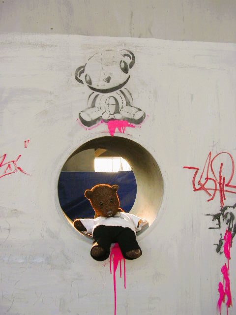 Bearsac sitting in round a hole in a wall with red paint that looks like blood. Above the hole isa garffitti teddy bear with a small bomb and blood under it.