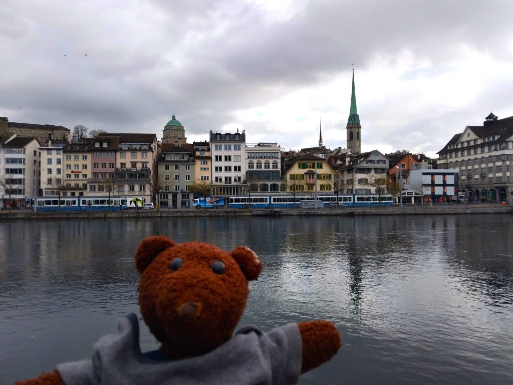 Bearsac the teddy bear in forground of a lake and buildings