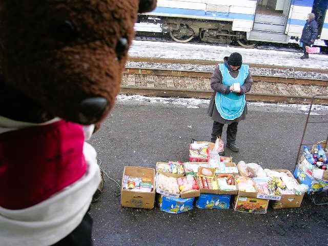 Bearsac the teddy bear in forgroud of a woman selling food item on a raliway platform.