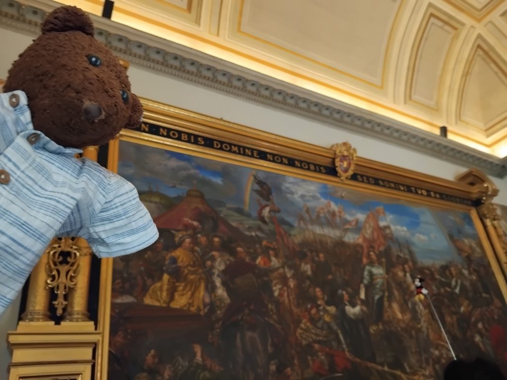 Bearsac the teddy bear in forground of a painign in the Vatican Museum.