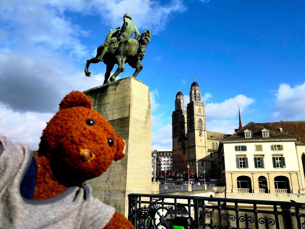 Bearsac the teddy bear in forground of a man and horse monument.