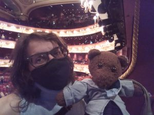 Debra wearing a face mask and holding Bearsac with an internal view of the Royal Opera House auditorium in the background.